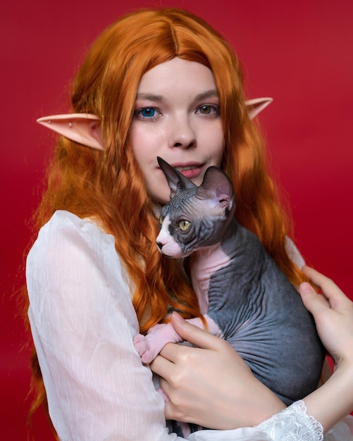 Portrait de jeune femme elfe cosplay tenant un adorable chaton Sphinx dans les mains sur fond rouge