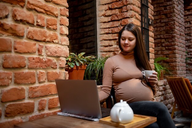 Portrait de jeune femme enceinte pigiste à l'aide d'un ordinateur portable pour un travail à distance tout en étant assis dans un café moderne avec une tasse de thé