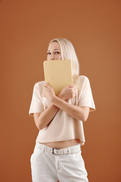 Portrait de jeune femme espiègle aux cheveux blonds cachant la bouche derrière la tablette sur fond marron