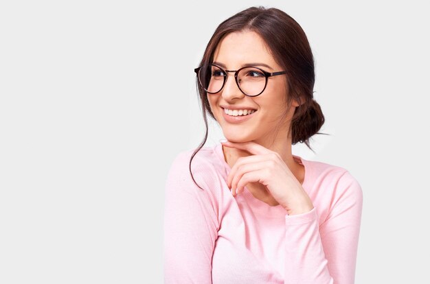 Portrait d'une jeune femme européenne assez positive en chemisier à manches longues rose et lunettes rondes transparentes souriant largement et regardant de côté isolé sur fond blanc Personnes de vraies émotions