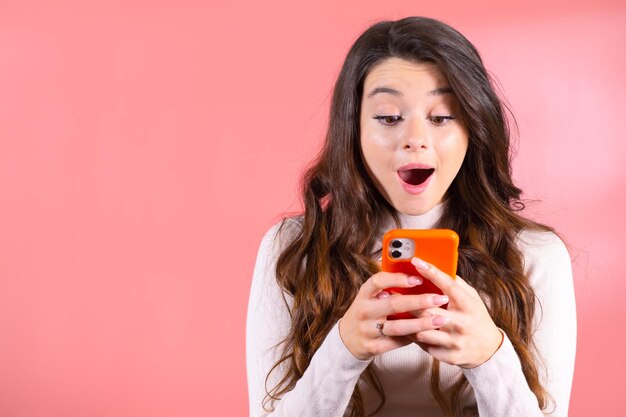 Portrait d'une jeune femme excitée faisant une surprise en regardant le téléphone portable isolé sur un fond rose