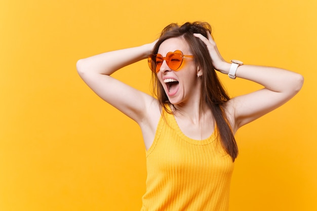 Portrait d'une jeune femme excitée qui crie dans des lunettes orange, met les mains sur la tête, garde la bouche grande ouverte, a l'air surpris isolé sur fond jaune. Concept d'émotions sincères de personnes. Espace publicitaire