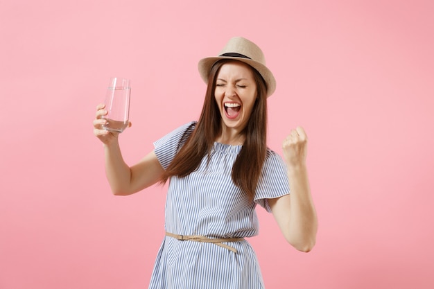 Portrait de jeune femme excitée en robe bleue, chapeau tenant et buvant de l'eau pure fraîche et claire à partir de verre isolé sur fond rose. Mode de vie sain, personnes, concept d'émotions sincères. Espace de copie.