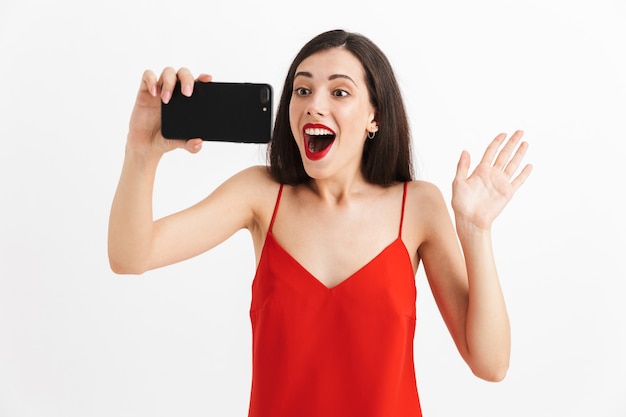 Portrait d'une jeune femme excitée en robe isolée, prenant selfie avec téléphone mobile, agitant