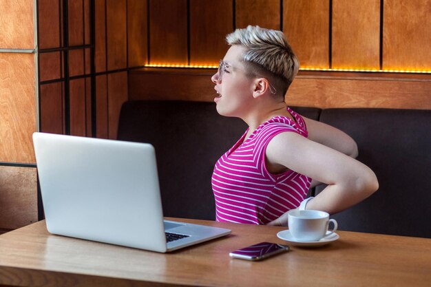 Portrait d'une jeune femme fatiguée aux cheveux blonds courts en t-shirt rose et lunettes assis dans un café et se tenant la main après une longue période assise sur l'ordinateur, faisant un massage. Intérieur, santé