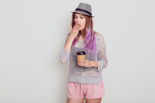 Portrait d'une jeune femme fatiguée portant un chapeau, ayant des cheveux violet vif, tenant une tasse jetable avec du café, a besoin d'énergie pour se réveiller, isolée sur fond gris.