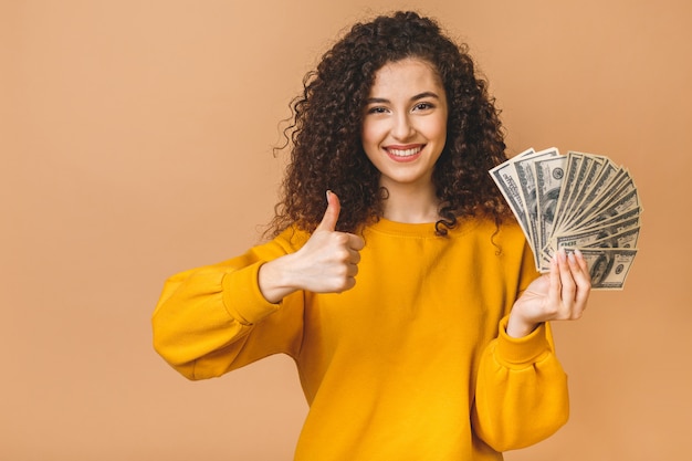 Portrait d'une jeune femme frisée gaie tenant des billets en argent et célébrant isolé sur fond beige. Pouces vers le haut.