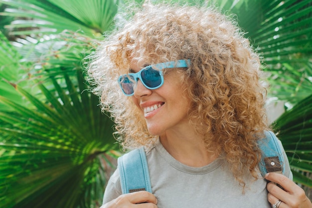 Portrait de jeune femme gaie adulte avec un sourire portant des lunettes de soleil bleues sur un fond vert tropical de feuilles Couleur et style de vie Cheveux longs bouclés blonds sains et bonheur Vie de voyage