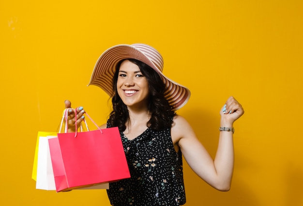 Portrait d'une jeune femme gaie avec chapeau tenant des sacs à provisions et présente des gestes isolés sur mur jaune