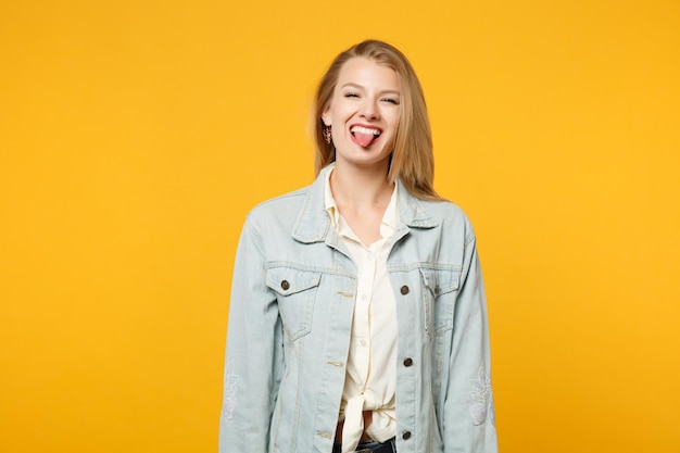 Portrait d'une jeune femme gaie dans des vêtements décontractés en denim regardant la caméra et montrant la langue isolée sur fond de mur orange jaune vif en studio. Concept de style de vie des gens. Maquette de l'espace de copie.