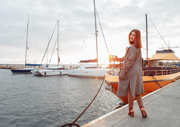 Portrait de jeune femme gaie en manteau d'automne posant dans un yacht club au lever du soleil