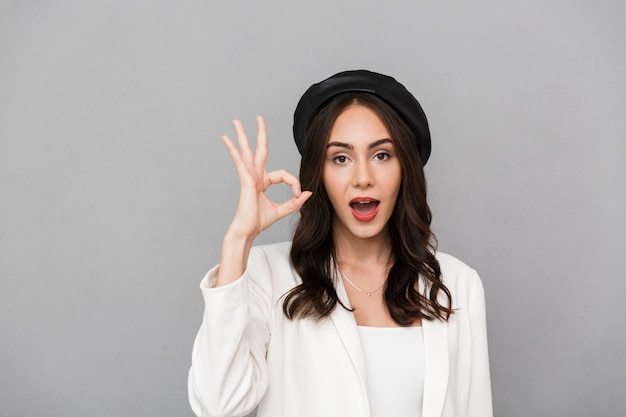 Portrait d'une jeune femme gaie portant béret debout isolé sur fond gris, montrant le geste ok