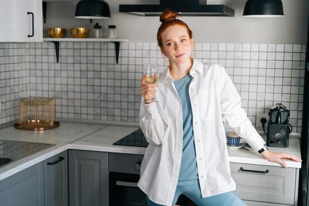 Portrait d'une jeune femme heureuse buvant du vin blanc debout dans une salle de cuisine lumineuse. Dame détendue dégustant du champagne seule tout en préparant la nourriture. Concept d'activité de loisirs femme rousse à la maison