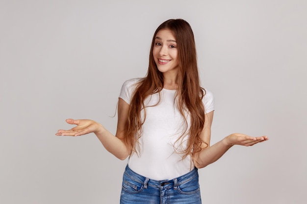 Portrait d'une jeune femme heureuse debout avec de larges bras levés et regardant la caméra avec un sourire à pleines dents partageant quelque chose, portant un T-shirt blanc. Tourné en studio intérieur isolé sur fond gris.