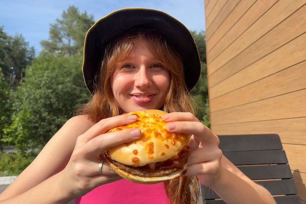 Portrait de jeune femme heureuse est en train de manger un délicieux burger juteux à l'extérieur du café en regardant la caméra