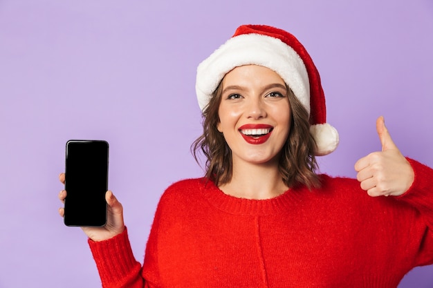 Portrait d'une jeune femme heureuse excitée portant un chapeau de Noël isolé sur un mur violet montrant l'affichage du téléphone mobile.
