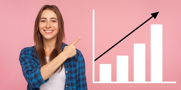 Portrait d'une jeune femme heureuse et joyeuse debout pointant de côté et montrant le graphique de croissance de l'entreprise prise de vue en studio à l'intérieur isolée sur fond rose