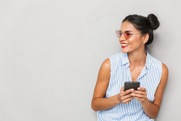 Portrait d'une jeune femme heureuse à lunettes de soleil isolé, à l'aide de téléphone mobile