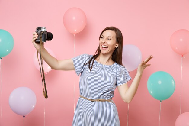 Portrait d'une jeune femme heureuse en robe bleue faisant du selfie sur un appareil photo vintage rétro écartant les mains sur fond rose avec des ballons à air colorés. Fête d'anniversaire personnes émotions sincères.