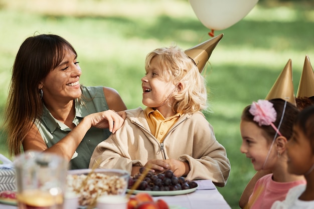 Portrait d'une jeune femme heureuse avec son fils assis à une table de pique-nique avec un groupe d'enfants pendant un bir en plein air...