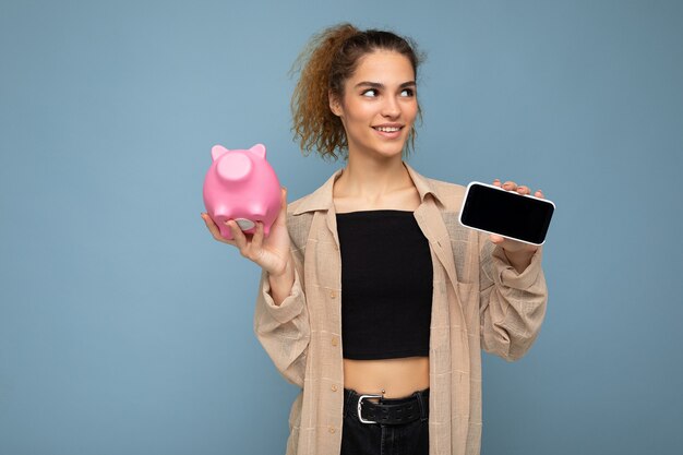 Portrait d'une jeune femme heureuse et souriante, séduisante et bouclée brune avec des émotions sincères portant une chemise beige élégante isolée sur fond bleu avec espace pour copie, détient de l'argent de cochon rose