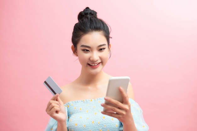 Portrait d'une jeune femme heureuse utilisant un téléphone portable, tenant une carte de crédit en plastique isolée sur fond rose