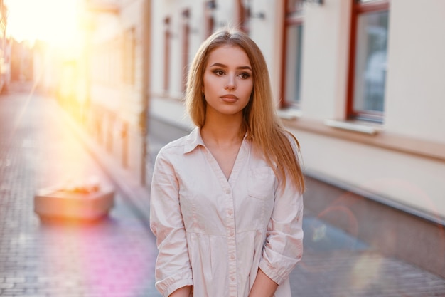 Portrait d'une jeune femme hipster aux beaux yeux bruns avec un maquillage naturel dans une élégante robe blanche d'été dans le contexte des rues de la ville et des couchers de soleil. Charmante fille en vacances