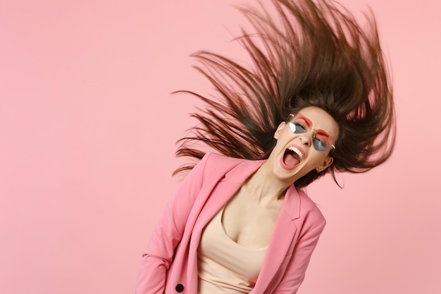 Portrait de jeune femme hurlant sauvage dans des lunettes de coeur sautant avec des cheveux qui coule isolé sur fond de mur rose pastel en studio. Émotions sincères des gens, concept de style de vie. Maquette de l'espace de copie.