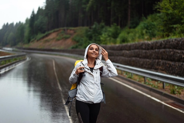 Portrait d'une jeune femme en imperméable debout sur une route asphaltée mouillée et souriante Charmant voyageur en randonnée dans les montagnes pendant les loisirs