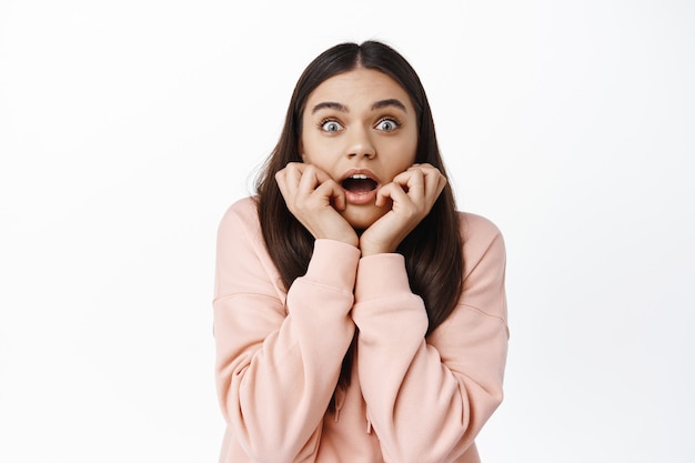 Portrait d'une jeune femme impressionnée haletant étonnée, tenant les mains sur le visage et bouche ouverte étonnée, vérifiant des nouvelles impressionnantes, regardant l'offre promotionnelle, mur blanc