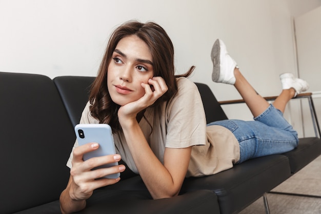 Portrait d'une jeune femme incroyable concentrée posant à l'intérieur à la maison sur un canapé à l'aide d'un téléphone portable.