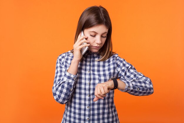 Portrait d'une jeune femme inquiète aux cheveux bruns portant une chemise à carreaux debout parlant sur un téléphone portable et regardant des montres sur son poignet, étant en retard. studio d'intérieur tourné isolé sur fond orange