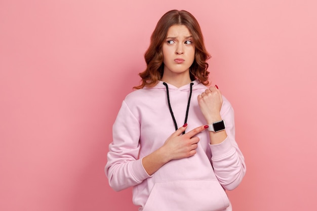 Portrait d'une jeune femme inquiète mécontente aux cheveux bouclés montrant une montre sur sa main rappelant l'heure tardive avertissant de la date limite Prise de vue en studio intérieure isolée sur fond rose