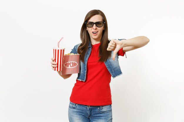 Portrait d'une jeune femme insatisfaite dans des lunettes 3d regardant un film, tenant un seau de pop-corn et une tasse en plastique de soda ou de cola, montrant le pouce vers le bas isolé sur fond blanc. Les émotions au cinéma.