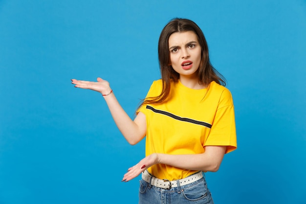 Portrait d'une jeune femme irritée et perplexe dans des vêtements décontractés vifs regardant la caméra, pointant les mains de côté isolées sur fond bleu vif en studio. Concept de style de vie des gens. Maquette de l'espace de copie.