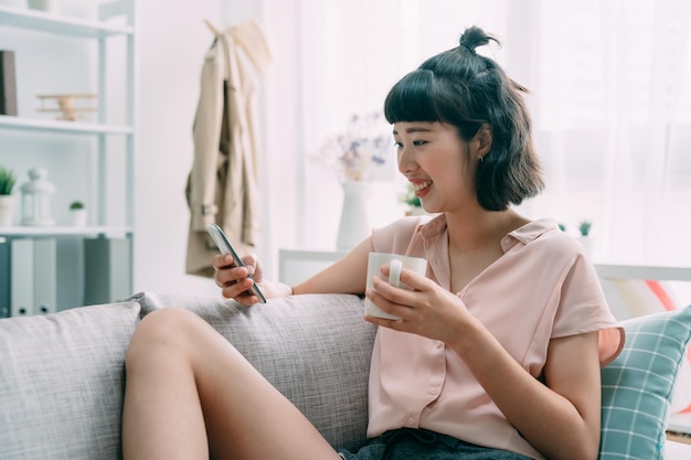 Portrait d'une jeune femme japonaise asiatique assise sur un canapé avec une tasse de café et un téléphone portable. fille heureuse en riant tout en discutant en ligne avec un ami sur un téléphone portable. les femmes apprécient la tasse de thé dans le canapé.