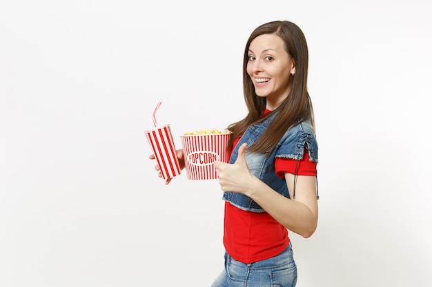 Portrait de jeune femme jolie souriante dans des vêtements décontractés en regardant un film, tenant un seau de pop-corn, une tasse en plastique de soda ou de cola montrant le pouce vers le haut isolé sur fond blanc. Les émotions au cinéma.