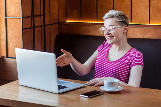 Portrait d'une jeune femme joyeuse aux cheveux courts en t-shirt rose et lunettes est assise sur un café et parle avec son amie sur un ordinateur portable et salue avec un sourire à pleines dents au mode de vie intérieur de la caméra Web