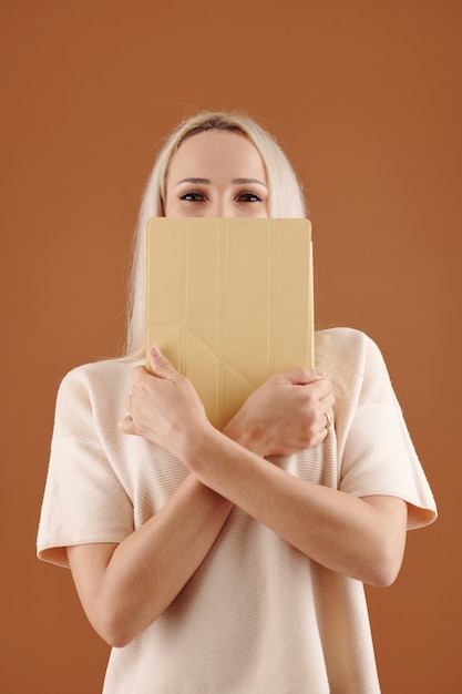 Portrait de jeune femme joyeuse couvrant le visage avec une tablette et regardant la caméra