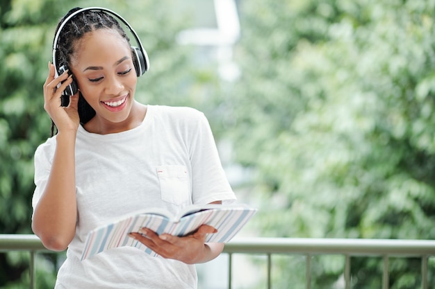 Portrait d'une jeune femme joyeuse dans des écouteurs lisant du texte dans un livre lors de l'apprentissage d'une autre langue