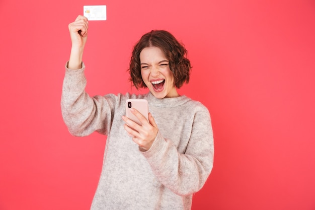 Portrait d'une jeune femme joyeuse debout isolé sur rose, à l'aide de téléphone mobile, montrant une carte de crédit en plastique