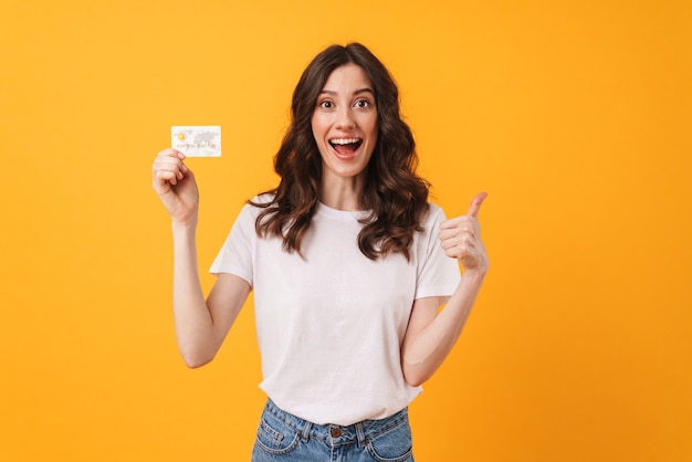 Portrait d'une jeune femme joyeuse et heureuse optimiste posant isolée sur un mur jaune tenant une carte de débit montrant les pouces vers le haut.