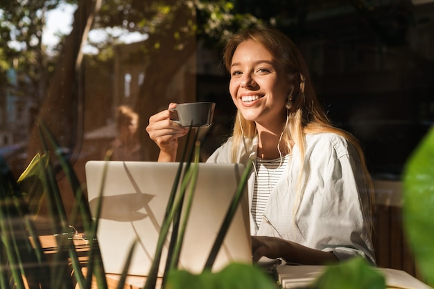 Portrait d'une jeune femme joyeuse portant des écouteurs buvant du café et utilisant un ordinateur portable assis au café