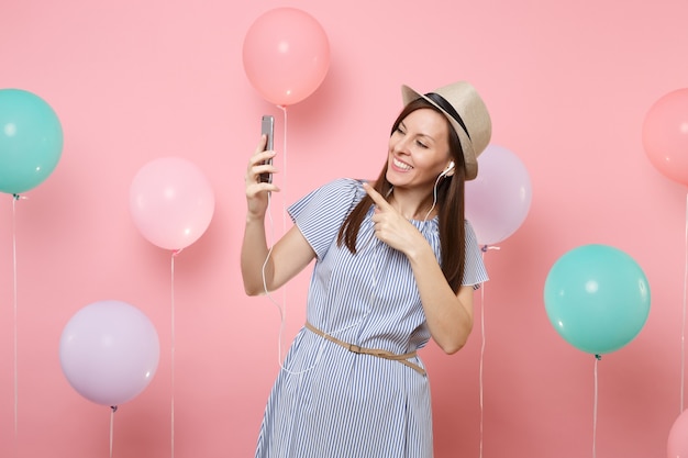 Portrait d'une jeune femme joyeuse en robe bleue de chapeau d'été de paille avec téléphone portable et écouteurs écoutant de la musique faisant un appel vidéo sur fond rose avec des ballons à air colorés. Fête d'anniversaire.