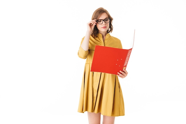 Portrait de jeune femme à lunettes et robe jaune debout et regardant pensivement dans un dossier rouge ouvert sur fond blanc isolé