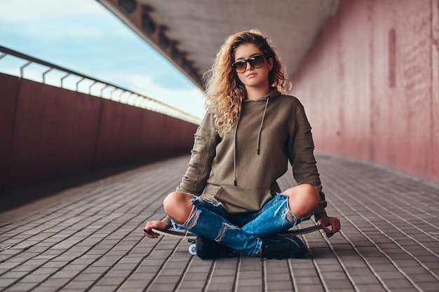 Portrait d'une jeune femme à lunettes de soleil vêtue d'un sweat à capuche et d'un jean déchiré assis sur une planche à roulettes avec les jambes croisées sur un trottoir de pont.