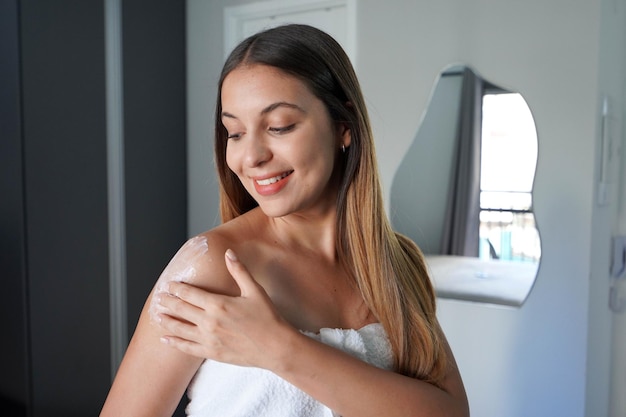 Portrait d'une jeune femme magnifique aux cheveux longs appliquant de la crème corporelle sur son épaule Lotion corporelle crème hydratante protection solaire concept de cosmétiques