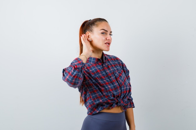 Portrait de jeune femme avec la main derrière l'oreille, debout sur le côté en chemise à carreaux et l'air curieux.