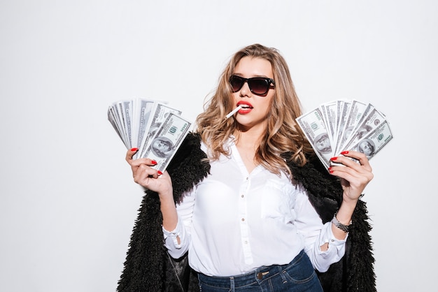 Photo portrait d'une jeune femme en manteau de fourrure tenant des billets d'un dollar américain et fumant une cigarette sur un mur blanc