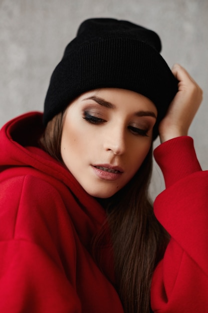 Portrait d'une jeune femme avec un maquillage lumineux dans un chapeau noir et un sweat à capuche rouge